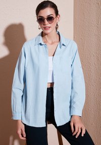 Camicia azzurra chiaro in un tessuto texturizzato, con tasca, maniche lunghe e orlo leggermente sfilacciato. Indossata con pantaloni neri.