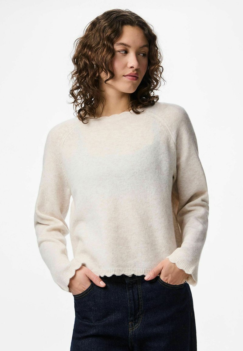 Jeune femme aux cheveux bouclés portant un pull crème à manches longues avec des bords festonnés, les mains dans les poches d'un jean bleu foncé, regardant vers la gauche.