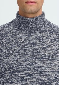 Moški nosi turtleneck pulover v mornarsko modri in beli melirani barvi, razkrije spodnji del obraza in vrat.