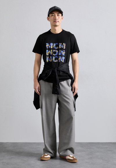 T-shirt en coton noir avec un graphique "NOUVEAU" coloré, pantalon large gris, chemise noire boutonnée nouée, mocassins marron avec des lacets blancs.