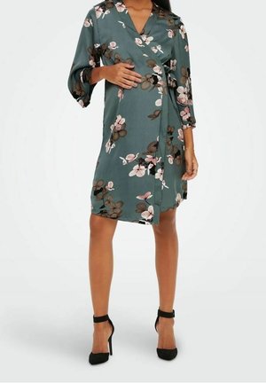 Woman wearing a green floral wrap dress with three-quarter sleeves and black ankle-strap high heels, standing against a plain background.