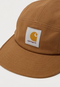 Καφέ καπέλο Carhartt από υφασμάτινη υφή με λευκή ορθογώνια κηλίδα λογότυπου που φέρει κίτρινο έμβλημα και κόκκινο κείμενο στο εμπρόσθιο πάνελ.