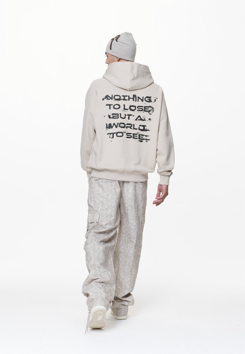 Beige Kapuzenpullover mit großem schwarzem Grafiktext auf dem Rücken, kombiniert mit hellgrauen gemusterten Cargo-Hosen und einer weichen grauen Mütze.
