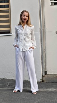 Blusa blanca de manga larga y bordados, combinada con pantalones de lino blancos holgados y sandalias planas blancas. Fondo neutro con un detalle met�álico.