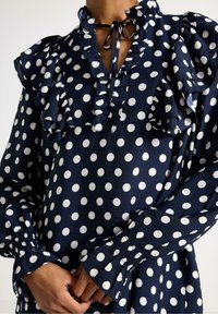 Mørkeblå bluse med hvide prikker, flæsekrave og mansjetter, snoretræk i halsen samt struktureret stof. Har lange ærmer og sammensynede detaljer.