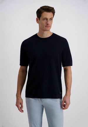 Man met kort bruin haar, gekleed in een donkerblauw shirt met korte mouwen en lichtblauwe broek, staand tegen een effen achtergrond.