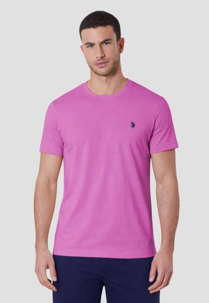 Uomo che indossa una maglietta rosa semplice a maniche corte con un piccolo logo scuro sul petto, in piedi davanti a uno sfondo chiaro e uniforme.