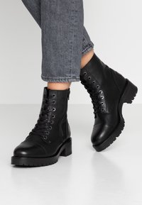 Zwarte leren enkellaarzen met veters en ritssluiting. Chunky hak en rubberen zool met profiel voor grip. Gestructureerde leren afwerking.