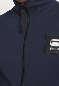 Marinblå zip-up jacka gjord av slätt material, med en svart logotypapplikation och en texturerad dragkedjehuvud vid kragen.