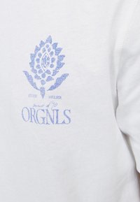 Camiseta de algodón blanca con un diseño gráfico azul de una planta estilizada y texto debajo. La tela parece suave con una textura lisa.