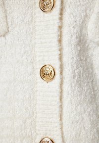 Vit stickad kofta med en texturerad yta, prydd med stora gyllene knappar med prägling. Tyget verkar mjukt och plyschigt.