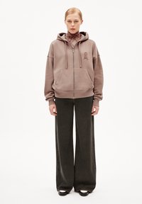 Haut à capuche zippé marron avec une poche avant, arborant un logo brodé discret. Associé à un jean ample noir et à des chaussures noires simples.