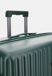 Grüner Hartschalenkoffer mit vertikal geripptem Design. Verfügt über einen grauen Teleskopgriff und eine Reißverschlussöffnung zur Aufbewahrung.