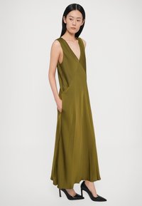 Olívazöld, ujjatlan, földig érő ruha mély V-nyakkivágással, átfedős dizájnnal és zsebekkel. Fekete magassarkú cipővel párosítva.