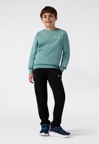 Sweatshirt bleu clair avec col rond et logo, associé à un pantalon de survêtement noir. La tenue est complétée par des chaussures sombres avec des accents bleus.