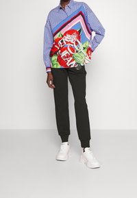 Vícebarevná nadměrná košile s modrými pruhy, květinovým vzorem a výrazným nápisem, doplněná o upnuté černé kalhoty a bílé tenisky.