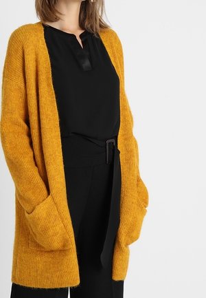 Femme portant un cardigan tricoté jaune moutarde par-dessus un chemisier noir et un pantalon noir ceinturé, mains dans les poches du cardigan.