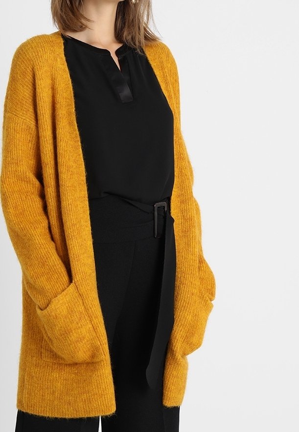 Femme portant un cardigan tricoté jaune moutarde par-dessus un chemisier noir et un pantalon noir ceinturé, mains dans les poches du cardigan.