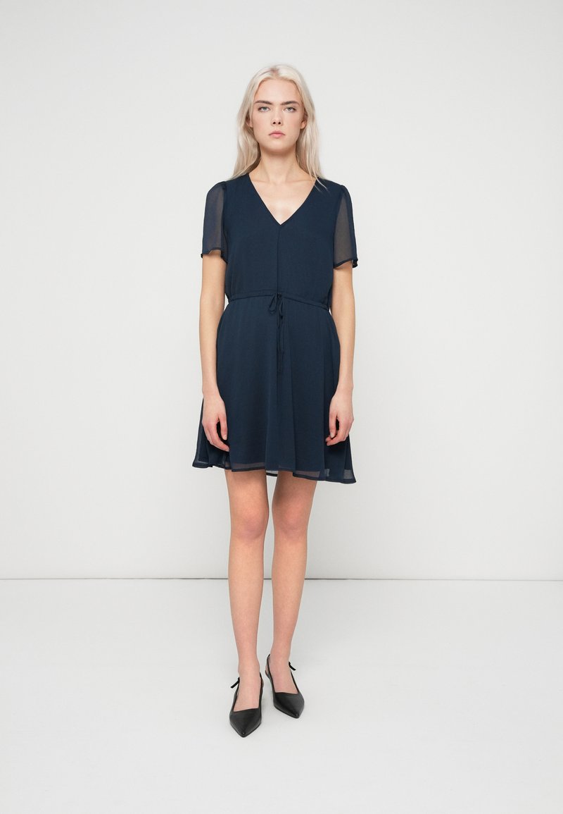 Γυναίκα με μακριά ξανθά μαλλιά, φορώντας ένα κοντό ναυτικό μπλε φόρεμα με διάφανες μανίκες και μαύρες ψηλοτάκουνες γόβες, που στέκεται μπροστά σε λευκό φόντο.