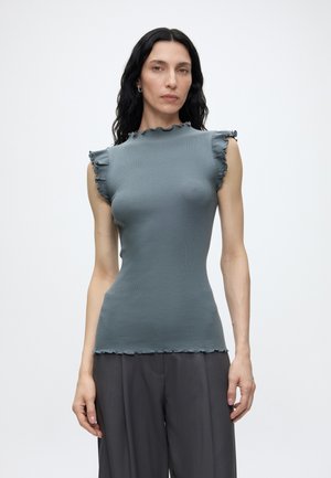 Femme aux longs cheveux foncés porte un haut gris ajusté sans manches avec des bords à volants et un pantalon ample gris foncé sur un fond clair uni.