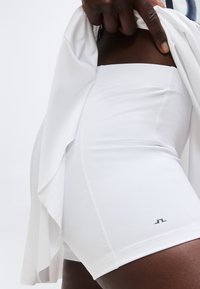 Detailný záber na osobu nosiacu biele športové šortky s vysokým pásom, ktorá zdvíha biely vrch, pričom sú viditeľné dolná časť trupu a horná časť stehien.