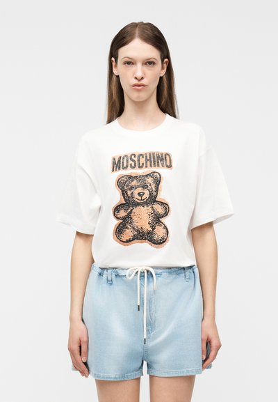 Γυναίκα που φοράει λευκό T-shirt Moschino με τυπωμένο αρκουδάκι και ανοιχτόχρωμα τζιν σορτς με κορδόνι, στέκεται μπροστά σε απλό φόντο.