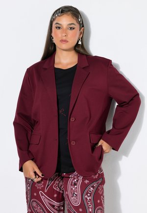 Kvinde med langt hår bærer sølv hårspænder, sølvøreringe, en mørkerød blazer, sort skjorte og mørkerøde bukser med paisley-mønster.