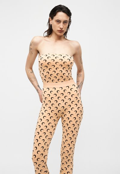 Oseba v breznaramničnem breskovem kombinezonu z vzorcem črnih polmesecov, pozira z rokami za hrbtom na enobarvnem ozadju.