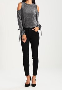 Svarta, kroppsnära jeans i kombination med en glittrig, silverfärgad topp med kalla axlar, prydd med knytningar vid ärmarna. Tydlig textur på materialet.