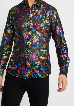 Ein Mann trägt ein tailliertes schwarzes Hemd mit einem lebhaften mehrfarbigen floralen Damastmuster und schwarze Hose vor einem schlichten Hintergrund.