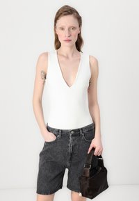 Body sans manches blanc avec un décolleté en V plongeant, associé à un short en denim noir taille haute et un sac à main compact marron foncé.