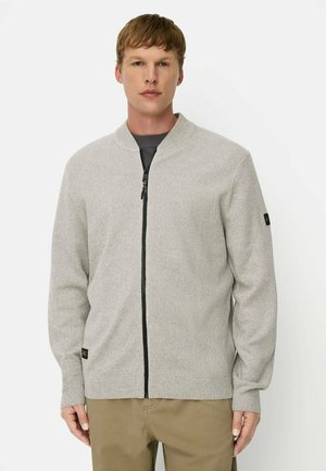 Man die een lichtgrijze sweater met zwarte rits draagt, beige broek en een donker overhemd eronder, staand tegen een witte achtergrond.