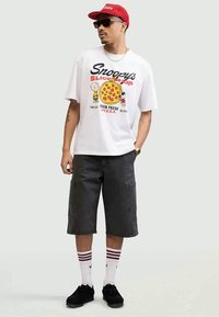 White graphic t-shirt with cartoon pizza design and characters, gray cargo shorts, red cap, and black sneakers with white-striped socks.