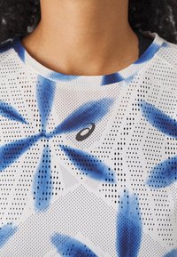 Short-sleeve athletic top in white with blue abstract floral patterns, featuring a round neckline and mesh texture, adorned with a small logo.
