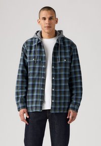 Plaid jacket in dark green, blue, and black tones. Features a grey hood, front snap buttons, and two chest pockets. Worn over a white shirt.