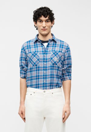 Młody mężczyzna o kręconych ciemnych włosach, ubrany w niebieską, kraciastą koszulę zapinaną na guziki z podwiniętymi rękawami oraz białe spodnie, stojący na jednolitym tle.