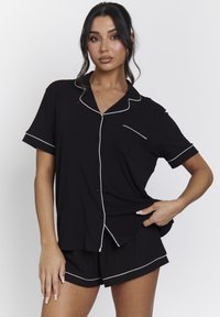Zwarte pyjama set met korte mouwen, bestaande uit een button-up shirt met een borstzak en witte piping, aangevuld met bijpassende shorts.