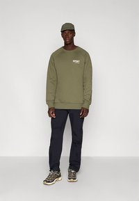 Õlivaheleroheline dressipluus logoga, mustad püksid ja hallid matkajalatsid. Mudel kannab ka sobivat mütsi, seistes ühevärvilise tausta ees.