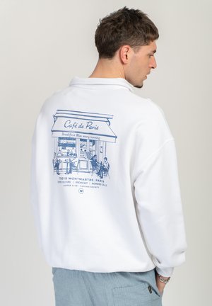 Mann trägt weißen Pullover mit blauem Entwurf des Café de Paris Schaufensters und sitzenden Personen auf der Rückseite, steht vor einfachem Hintergrund.