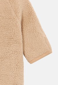 Beige, langermet, teksturert sherpa-fleece stoff med ribbestrikket mansjett på ermene, vist mot en hvit bakgrunn.