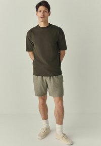 Donkergroen T-shirt met korte mouwen en lichtgrijze geruite shorts, gecombineerd met crèmekleurige sneakers en witte sokken, staand op een effen achtergrond.