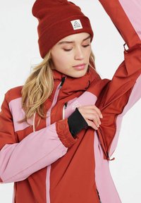 Rode en roze buitenjas met een rits, verstelbare manchetten en een mesh binnenzak; gedragen met een bijpassende rode gebreide beanie.