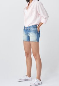Femme debout portant une chemise rose clair, un débardeur blanc, un short en jean bleu et des tennis en toile blanches, main dans la poche, sur un fond uni.