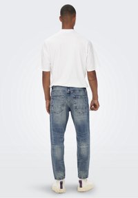 Valkoinen ylipitkä t-paita yhdistettynä sinisiin farkkuihin. Farkuissa on kulutettu viimeistely ja takataskut; asukokonaisuuteen kuuluu valkoiset lenkkarit.