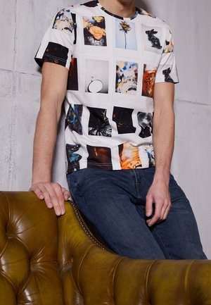 Man wearing white T-shirt with colorful photographic print squares, blue jeans, leaning on olive-green tufted leather chair.