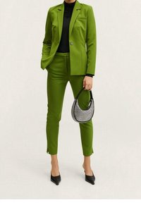 Personne portant un costume vert sur mesure avec un haut noir, des chaussures noires pointues et tenant un petit sac à main noir et blanc à motif.