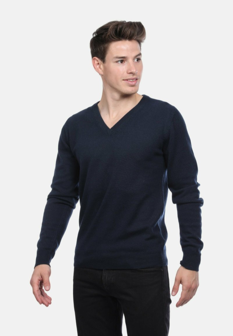 Un jeune homme portant un pull en V bleu marine et un pantalon noir, regardant légèrement vers la droite avec une posture détendue contre un fond blanc.