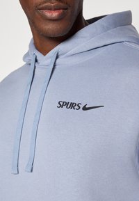 Svetlo modrá bavlnená mikina s kapucňou, s predným klokankovým vreckom, čiernym nápisom "SPURS" a logom Nike. Obsahuje kapucňu so šnúrkami a mäkkú textúru.