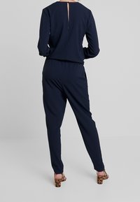 Combinaison marine à manches longues avec un détail en ouverture à l'arrière. Le tissu semble lisse avec une taille élastique. Portée avec des chaussures à talons hauts à motifs.