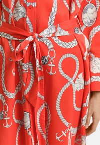Abito di stoffa rosso con motivo di corde e ancore, caratterizzato da una vita legata e una chiusura frontale con bottoni. Texture liscia e design fluido.